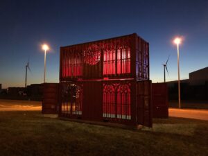 Een foto van de containerkathedraal bij nacht met twee verlichte lantaarnpalen.