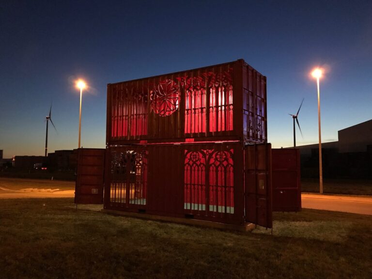 Een foto van de containerkathedraal bij nacht met twee verlichte lantaarnpalen.