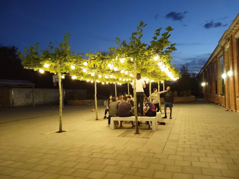 Groep leerlingen secundair onderwijs zit ‘s avonds op de binnenplaats van het jeugdcentrum op de banken onder de leibomen. © Havenwereld
