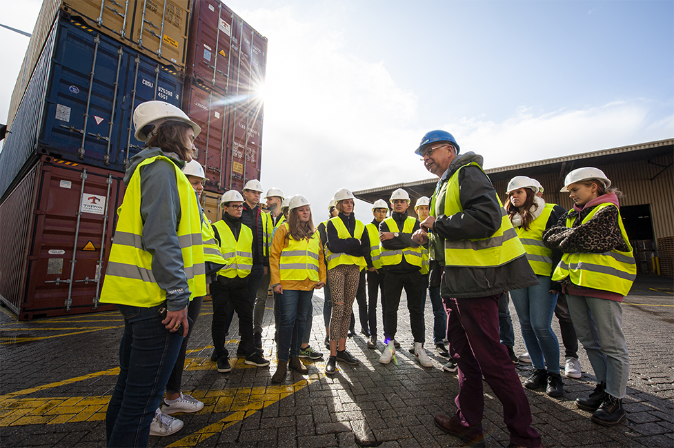 Cursisten volwassenenonderwijs luisteren naar uitleg over containerlogistiek naast gestapelde zeecontainers tijdens een bedrijfsbezoek in de haven. © Kris Jacobs