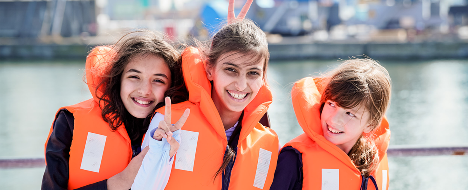 Drie meisjes van de derde graad lager onderwijs staan op het buitendek tijdens hun havenrondvaart, glimlachen en maken een v-teken met hun vingers. © Provincie Antwerpen