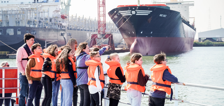 Een groepje leerlingen van de derde graad lager onderwijs staat buiten op het dek van het schip met een begeleider en gids. Ze varen langs enkele grote schepen tijdens een havenrondvaart. © Provincie Antwerpen