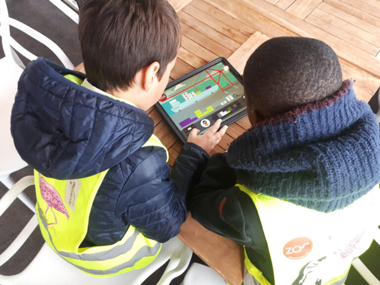 Twee kinderen van het lager onderwijs in veiligheidsvesten werken samen aan het lespakket op een tablet aan een houten tafel. © Havenwereld