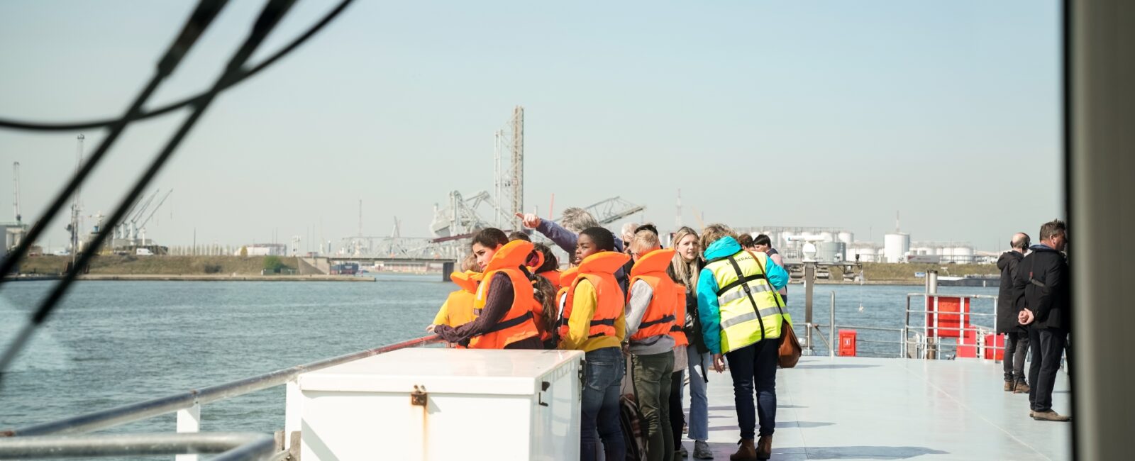 Een groep leerlingen van de derde graad lager onderwijs staat buiten op het dek van het schip met hun begeleider tijdens een havenrondvaart. Een jongen wijst naar iets dat hij gezien heeft in de omgeving. © Provincie Antwerpen