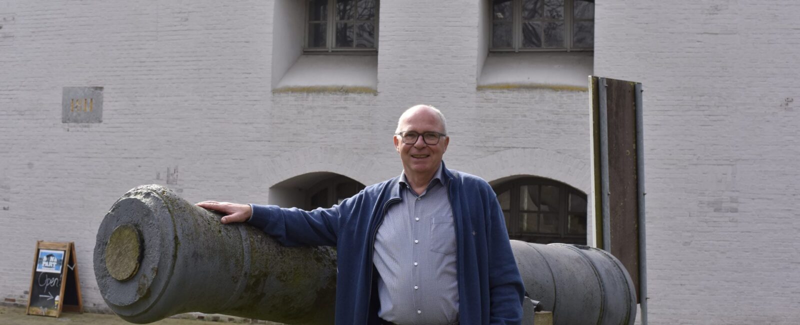 Man met blauwe cardigan staat voor een oude kanon