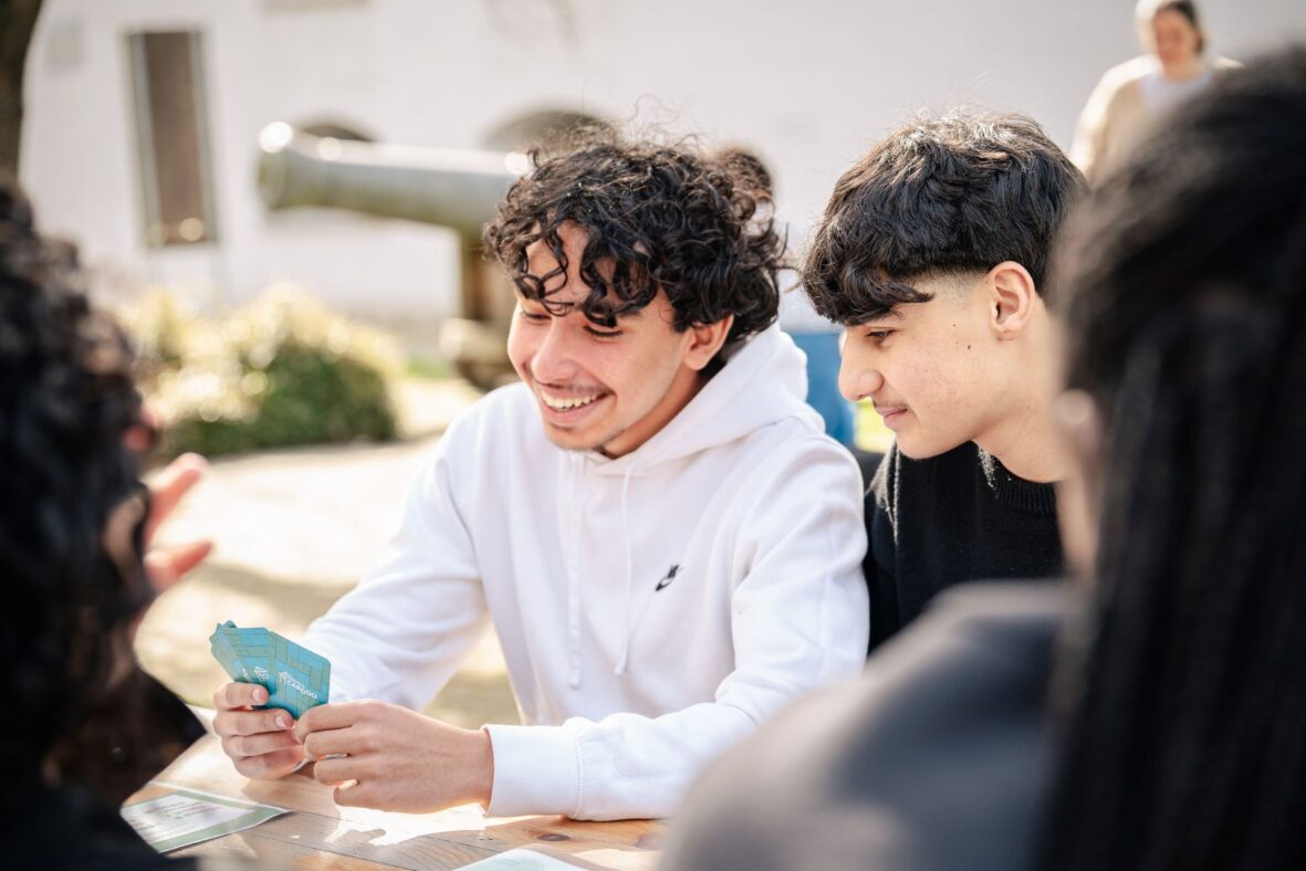 Twee jongens van de derde graad secundair onderwijs bekijken samen de set Cardgo-spelkaarten die de ene jongen in de hand heeft. Hij glimlacht breed. © Tom Cornille