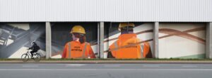 afbeelding van twee havenarbeiders die in rugaanzicht op een muur geschilderd staan, terwijl er een fietser voorbijrijdt