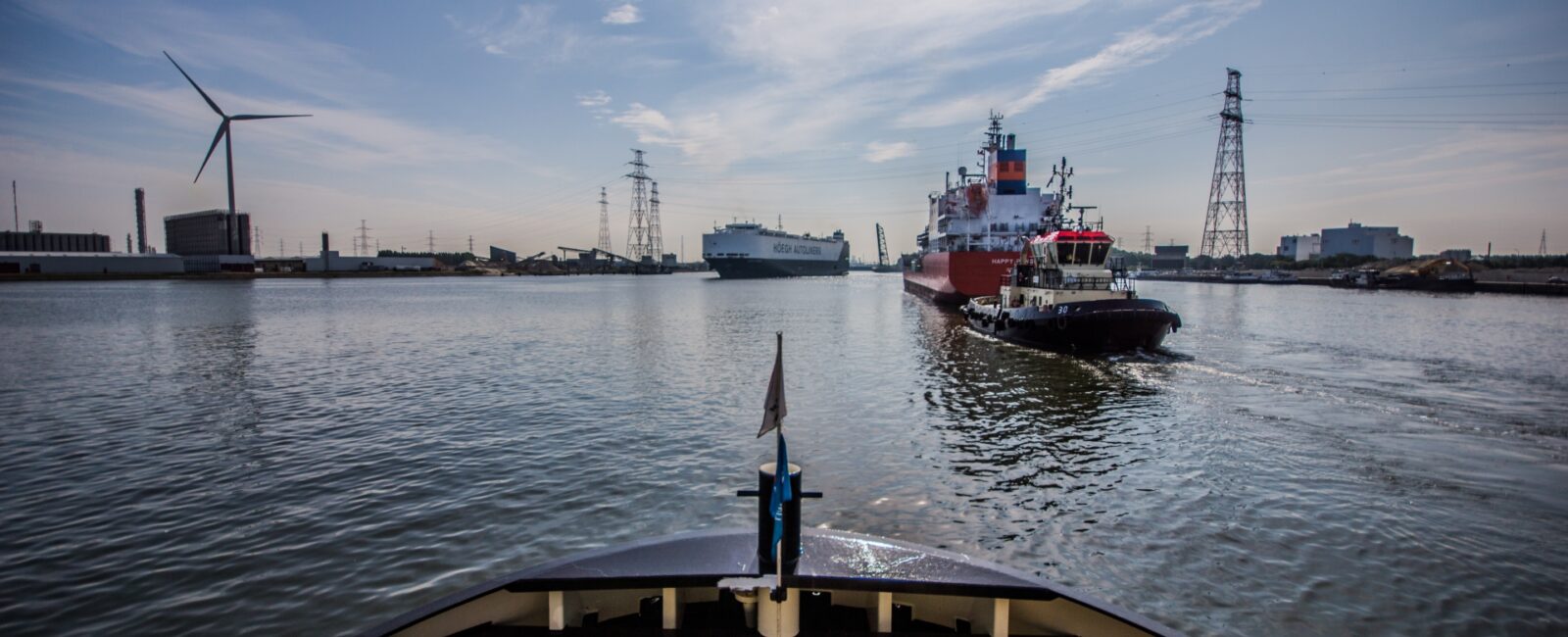 Beeld vanop een schip in de haven. We zien andere schepen, industriegebouwen, hoogspanning en een windmolen. © Den Antwerpenaar